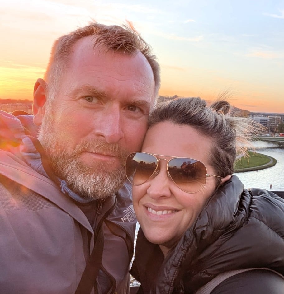 A person wearing a North Face jacket and another person with sunglasses are embracing, with a scenic river and cityscape at sunset in the background.