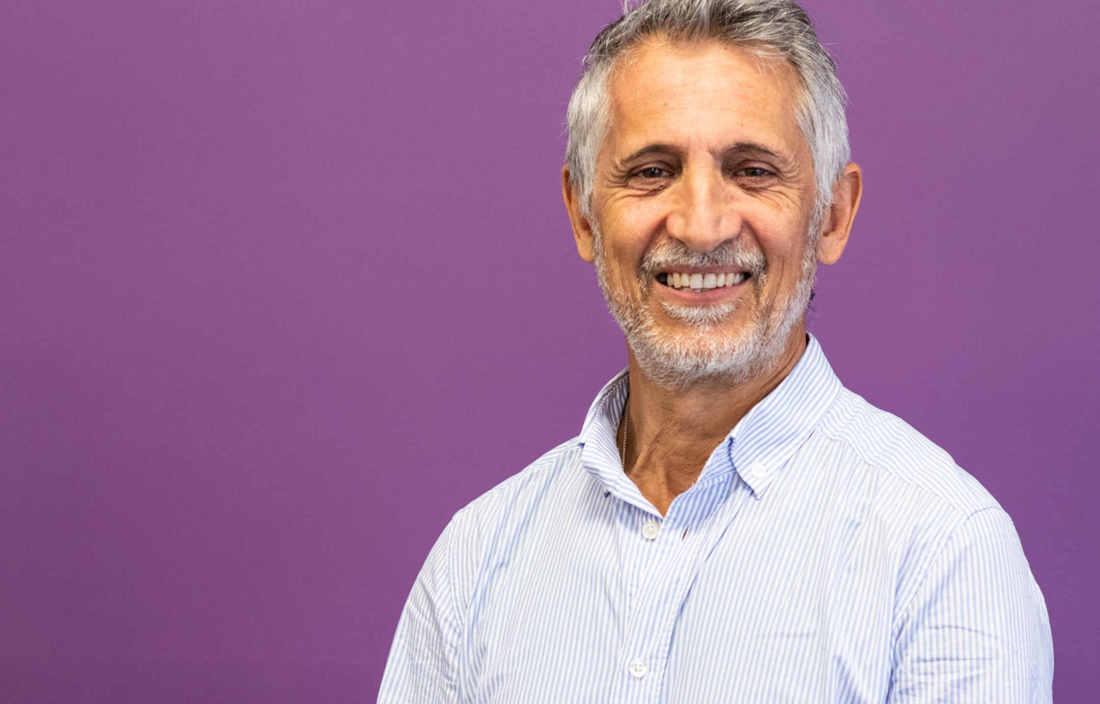 Saeed - a man in his 70s with grey hair and light beard wears a white shirt and is photographed against a purple background
