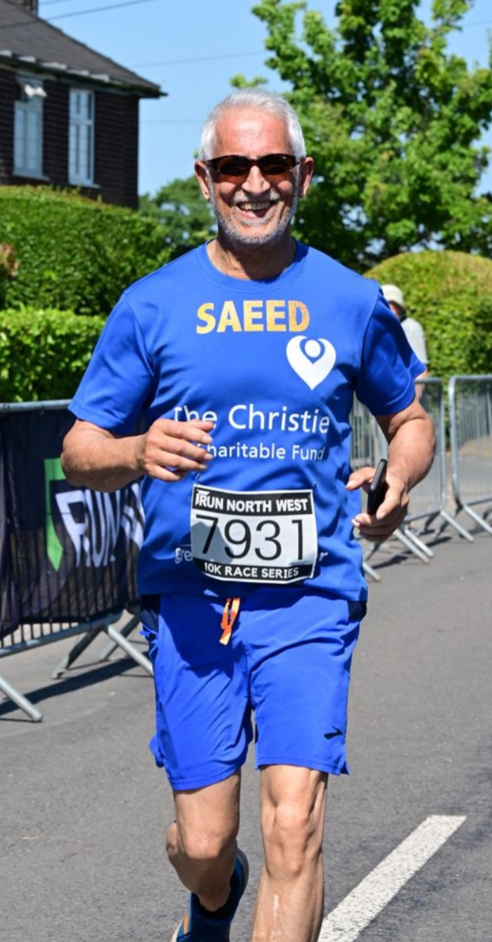 Saeed - a man in his 70s with grey hair and beard wear sun glasses and blue shorts and a T-shirt with The Christie written on it as he runs in a marathon