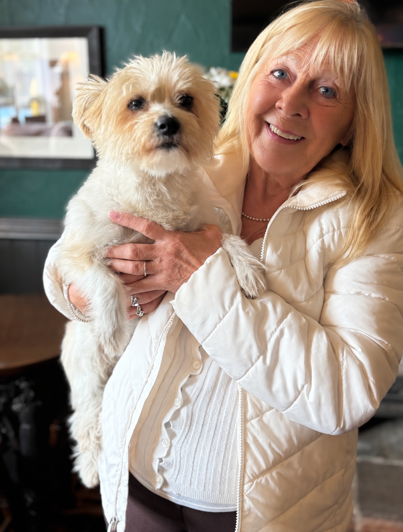 Jean Page - a white lady in her 70s with long blonde hair stands indoors holding her dog Dolly.