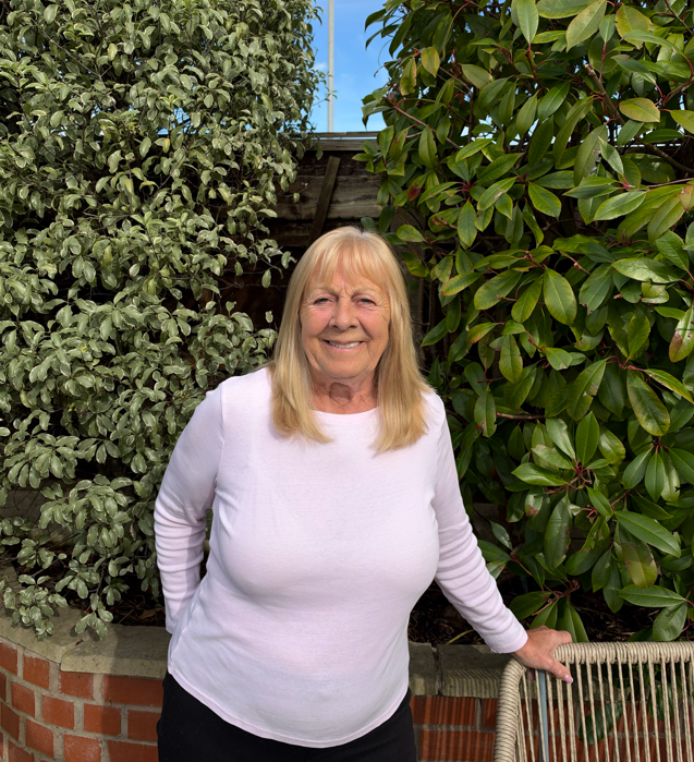 Jean Page - a white lady in her 70s with long blonde hair stands in front of the green hedge outside