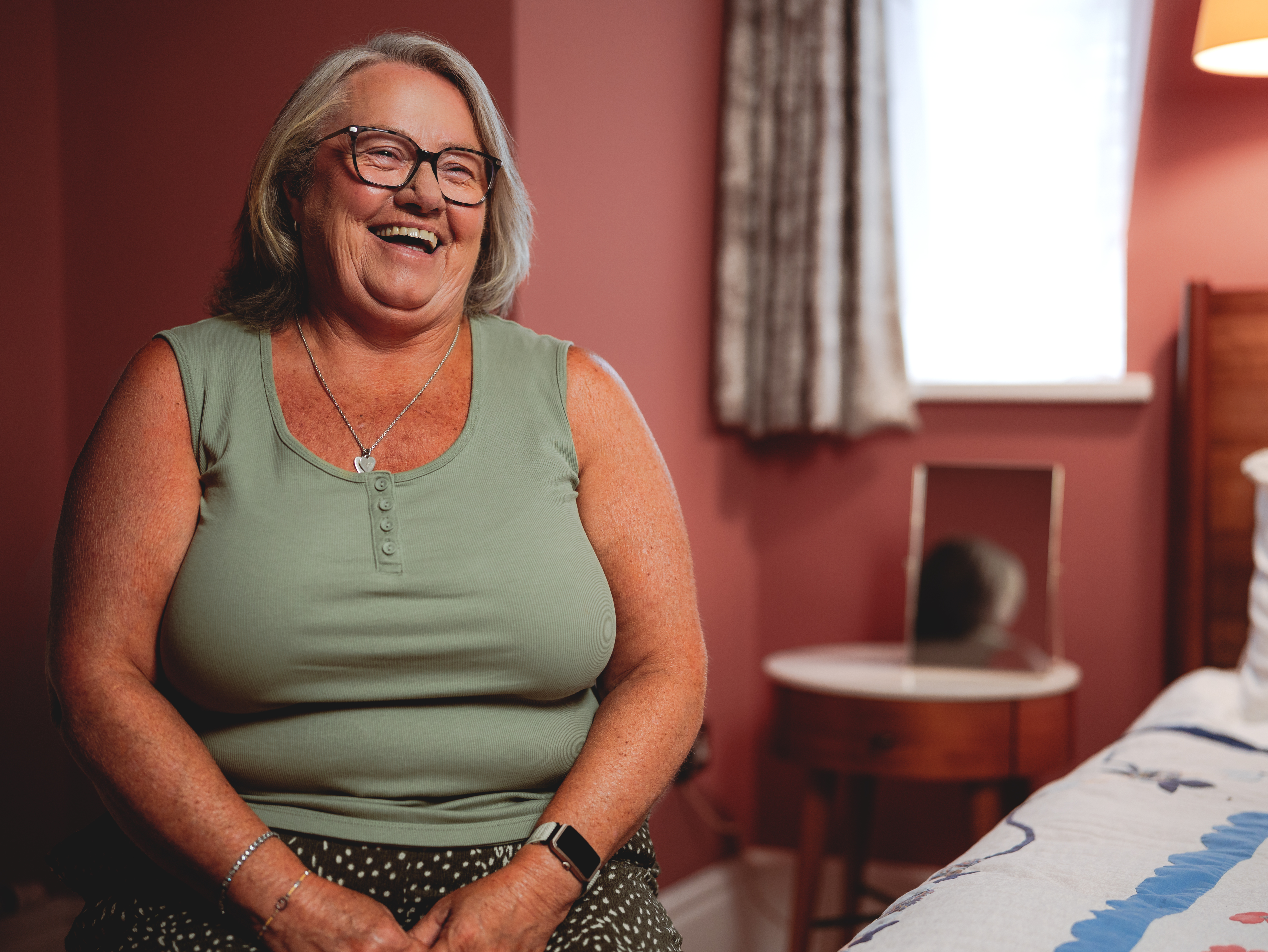 Gillian a white woman in her 60s with chin length grey hair and glasses sits indoors and smiles at the camera 