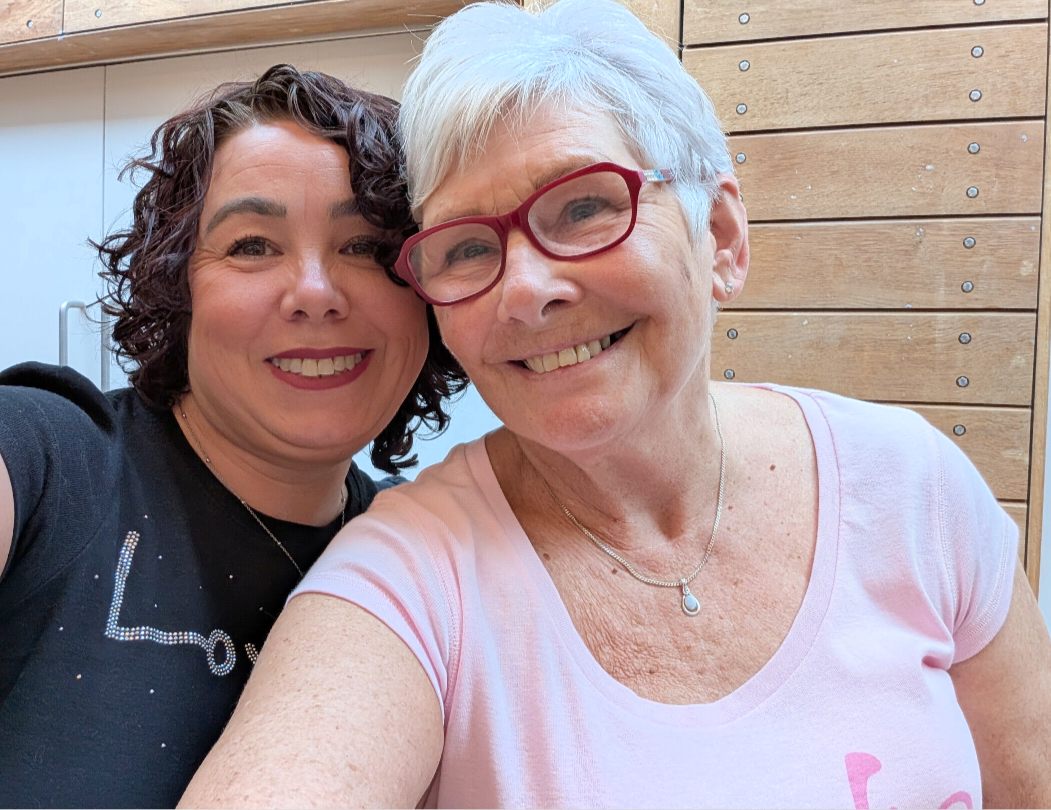 Lindsey a woman with dark curly hair in her 40s with Janet a woman with short grey hair and glasses in her 70s indoors posing for a selfie