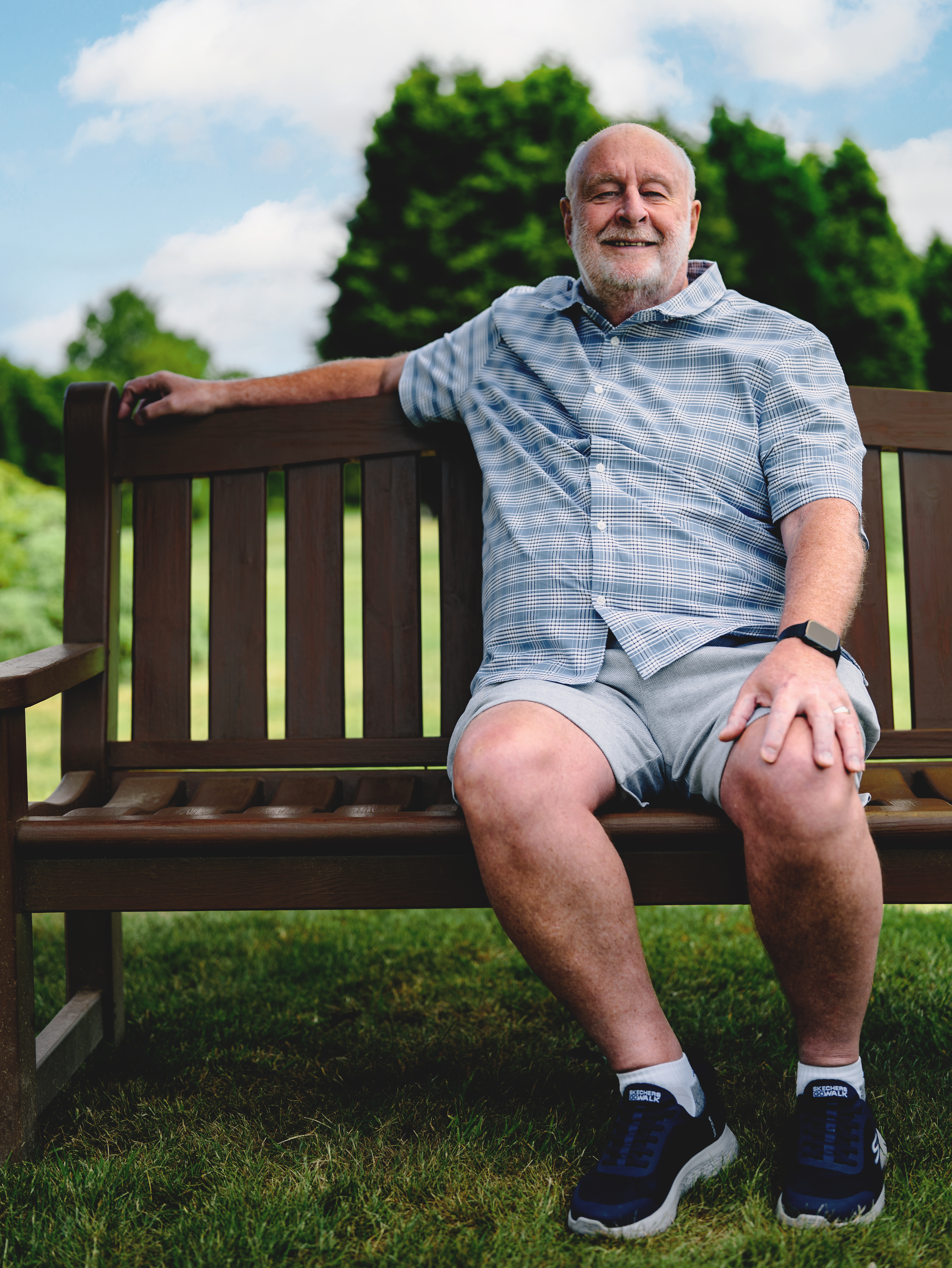 Ken a man with grey hair and a beard sits on a bench in a park wearing a checked shirt and shorts