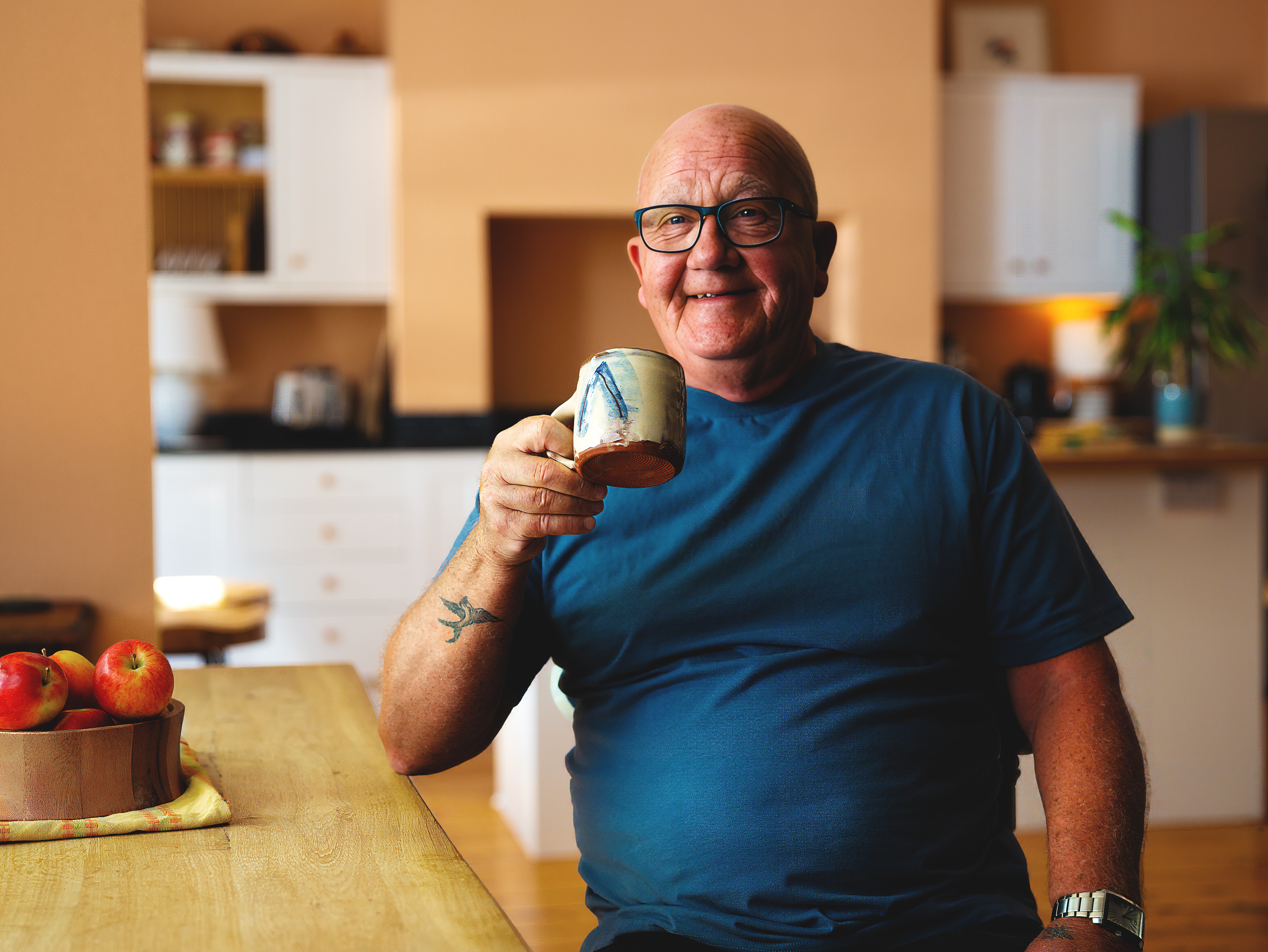 Phil a white man in his mid-60s with glasses and a bald head sits with a mug of tea in a kitchen